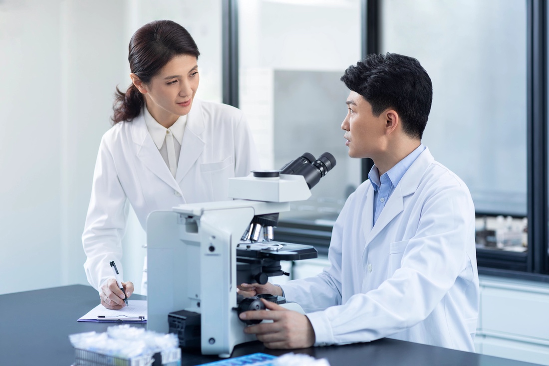 Two people in lab coats - one at microscope, one taking notes