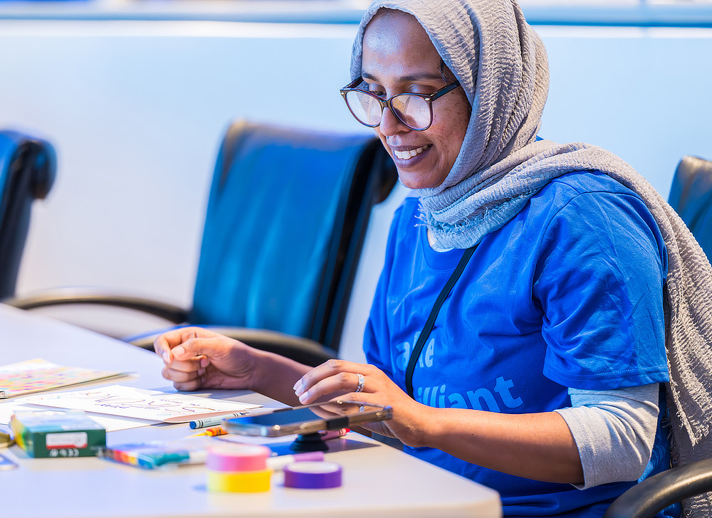 A woman wearing a headscarf and glasses is coloring a picture at a table. She is smiling and appears to be enjoying the activity.