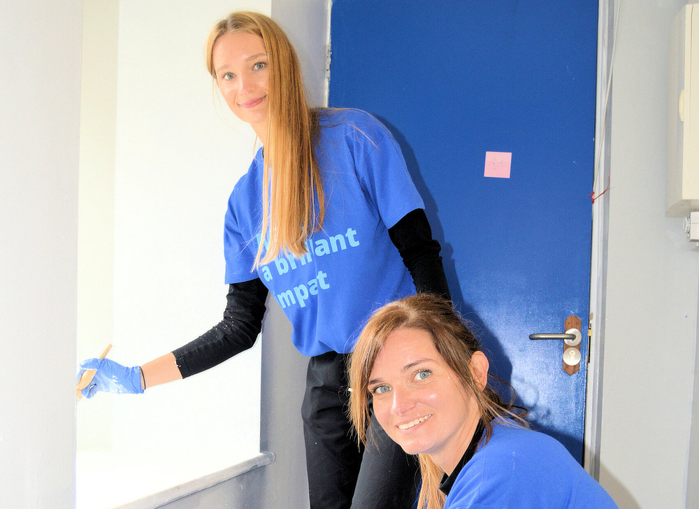 Two women are painting a wall white. They are wearing blue shirts and gloves, and one is kneeling while the other stands.