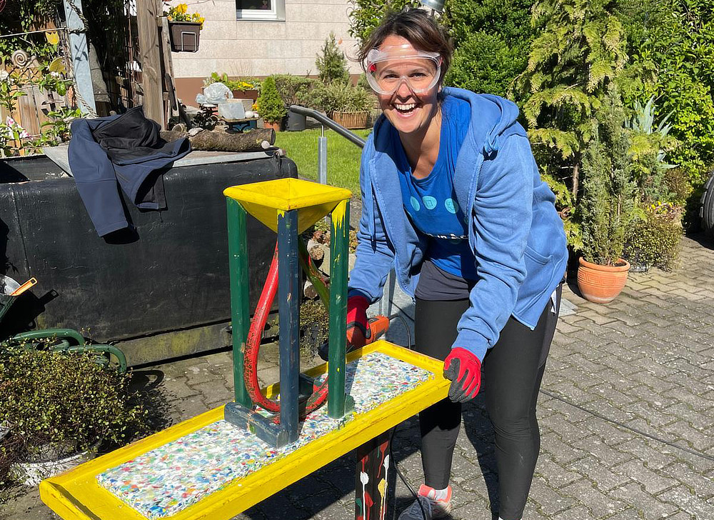 A woman is working on a brightly colored table in a garden. She is wearing gloves and safety glasses, and appears to be applying something to the tabletop.