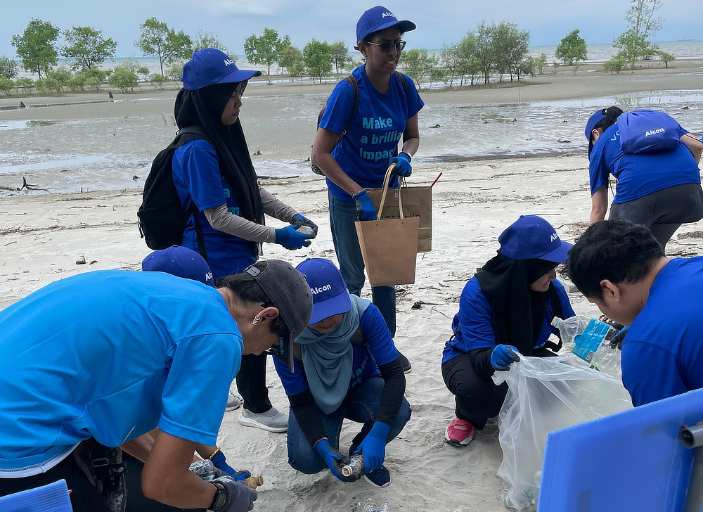 A group of volunteers wearing blue shirts are picking up trash on a muddy beach. They are collecting plastic bottles and other debris.
