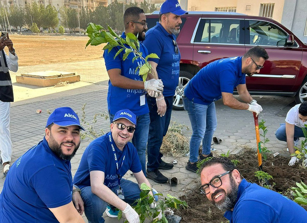 A group of volunteers wearing blue shirts are planting small trees in a garden bed. They are smiling and working together.