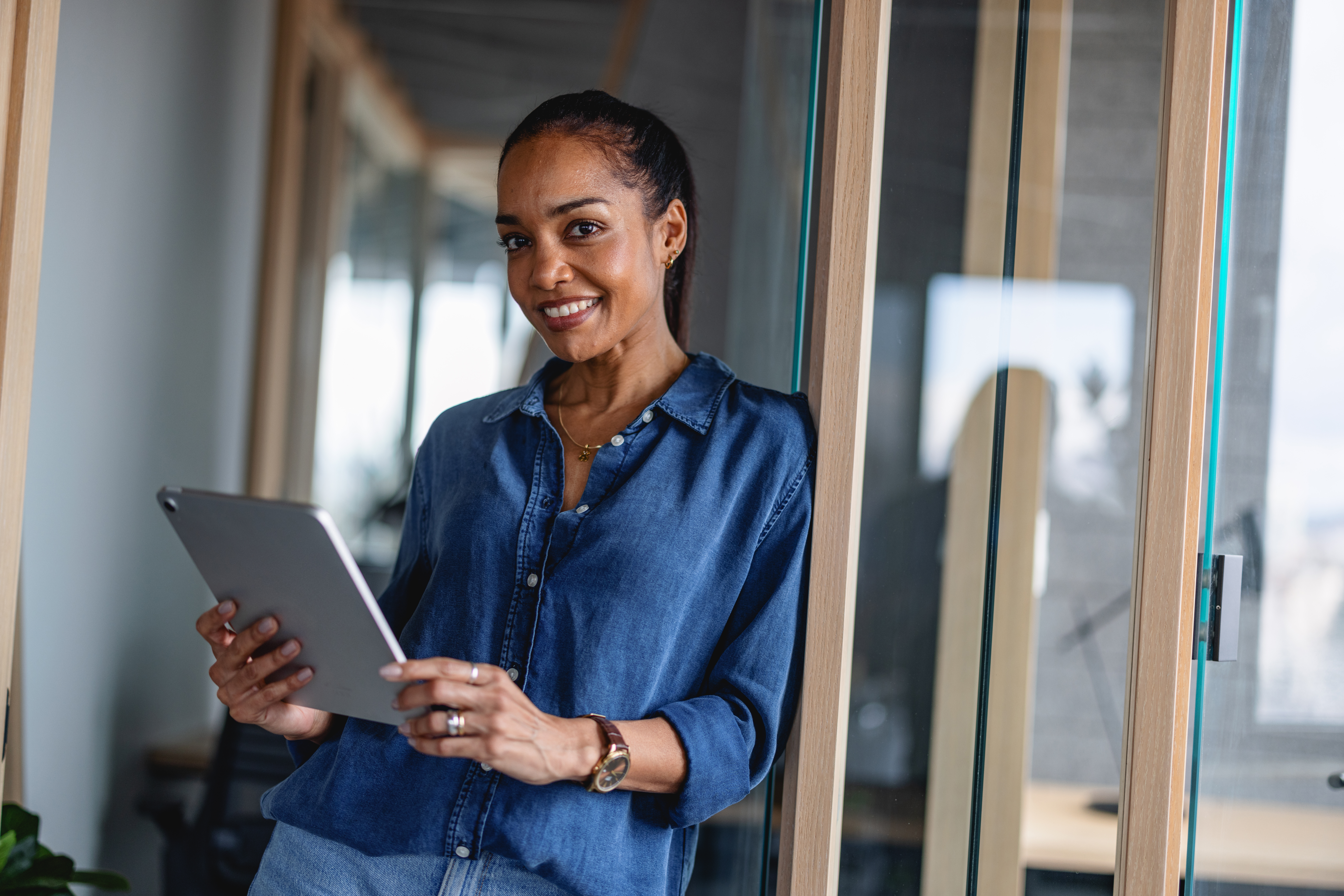 A smiling woman is looking down at a tablet she is holding in both hands. She is wearing a blue button-down shirt and a watch.