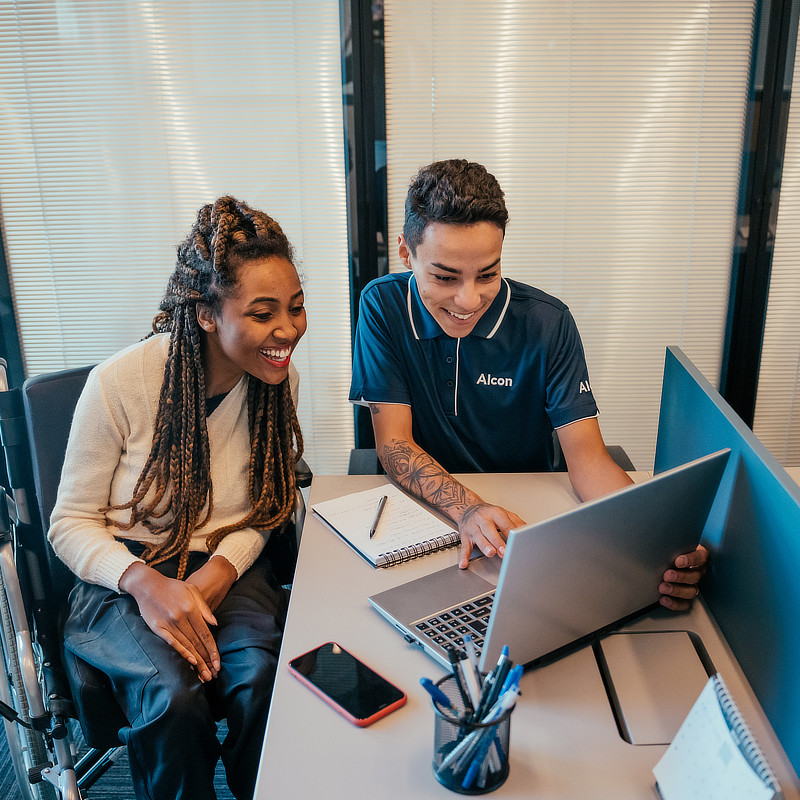 Two colleagues are leaning over a laptop at a desk in an office setting. They appear to be collaborating. One person is wearing a white sweater and the other is wearing a navy polo shirt with an Alcon logo on it.