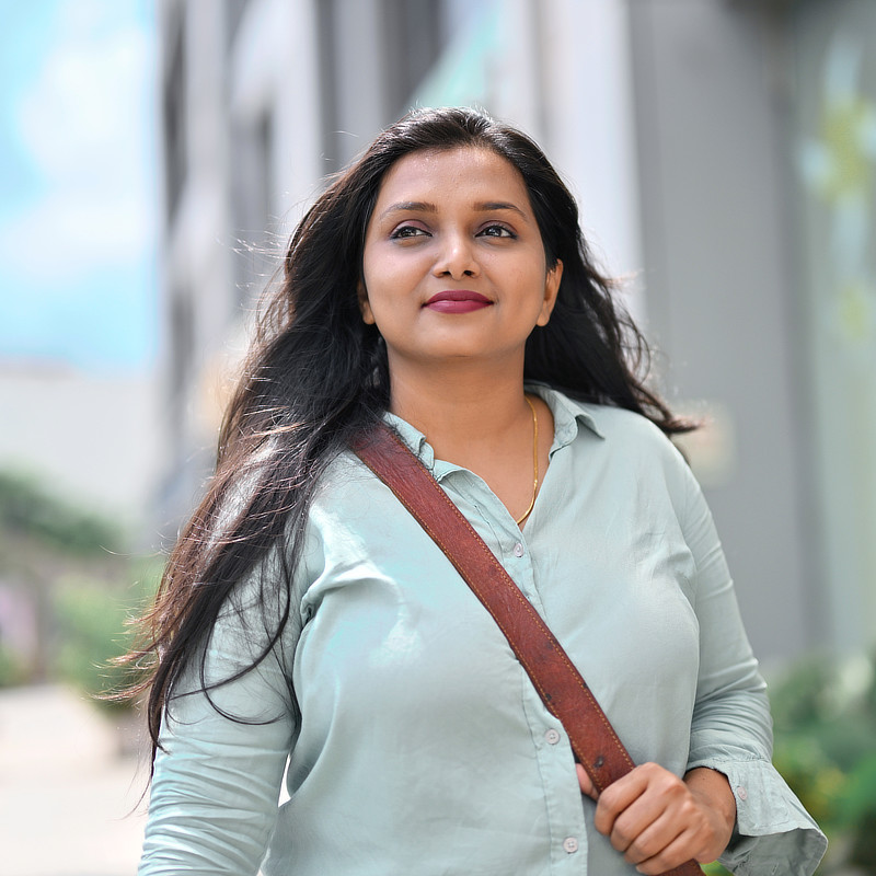 A woman with long, dark hair and brown eyes looks directly at the camera. She is wearing a light blue button-down shirt with a dark brown strap bag slung over her shoulder.