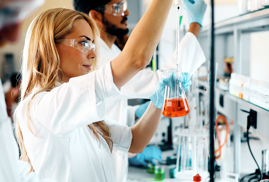 A female scientist wearing a lab coat and safety glasses carefully examines a flask containing an orange liquid.