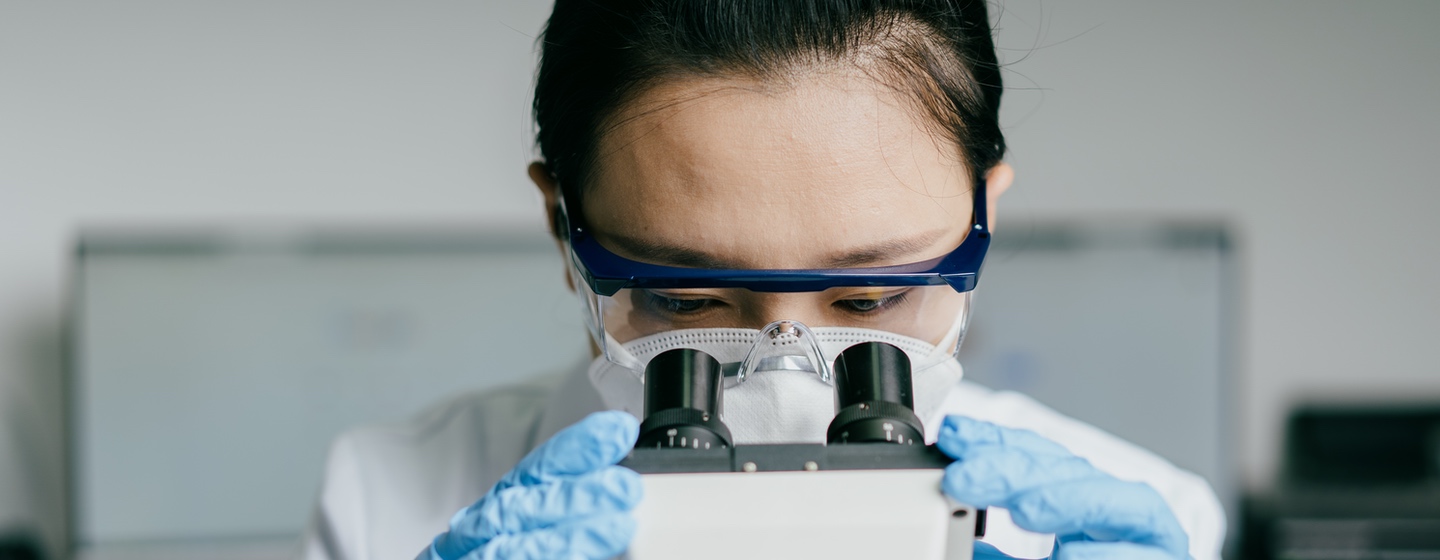Woman in lab coat and gloves looking through microscope